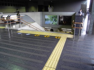 Colour photograph showing a main circulation route at a Railway Station in Kyoto, Japan ... with combined staircase and elevators. Notice, in particular, the dual height staircase handrails, for adults and children ... the strong contrast of the floor tactile information (a 'directional' indicator leading to a 'hazard' indicator, at the top of the staircase) compared to the rest of the floor, with its broad non-slip strips ... and, finally, arrows used to control staircase circulation flows at peak periods (down to the right, up on the left). Photograph taken by CJ Walsh. 2010-04-27. Click to enlarge.