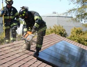 Colour photograph showing two firefighters on a roof, one with cutting equipment. Solar Photovoltaic Roof Panels restrict firefighter access to building interior roof spaces.